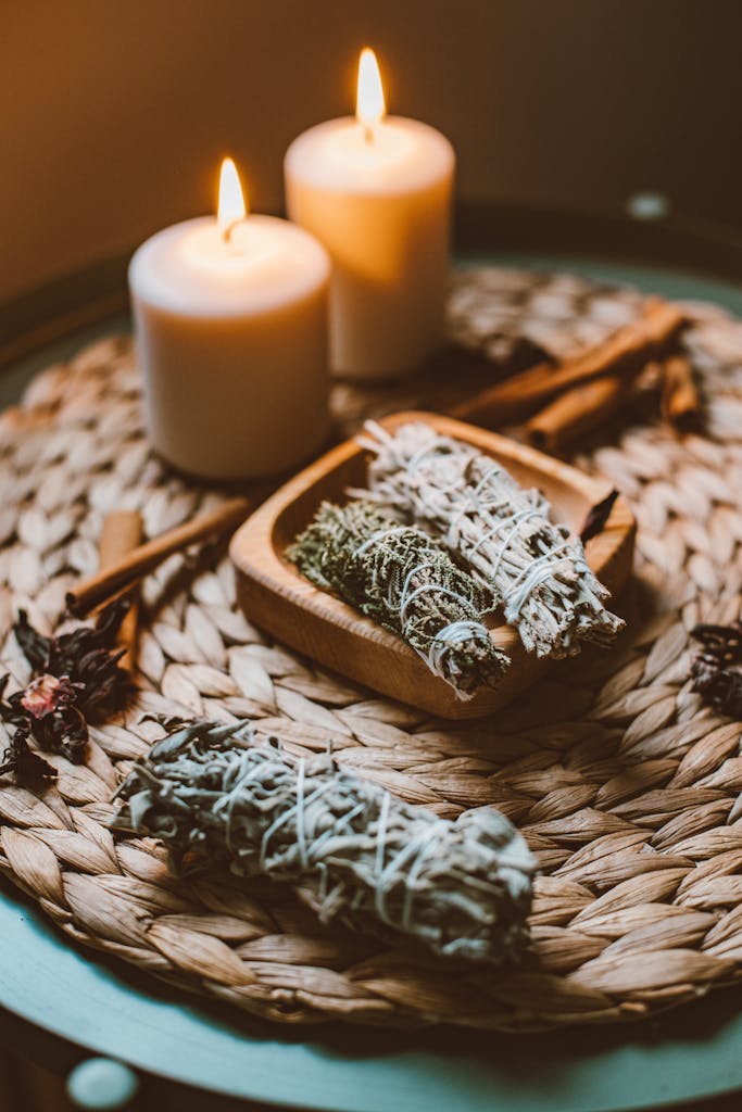 A warm and inviting scene with candles, sage bundles, and cinnamon sticks on a woven mat.